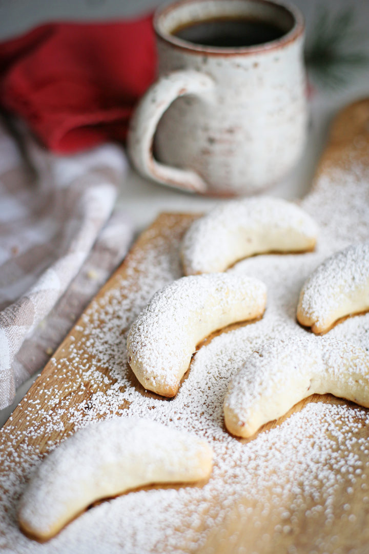 photo of a batch of almond crescent cookies being dusted with powdered sugar on a rustic wooden cutting board