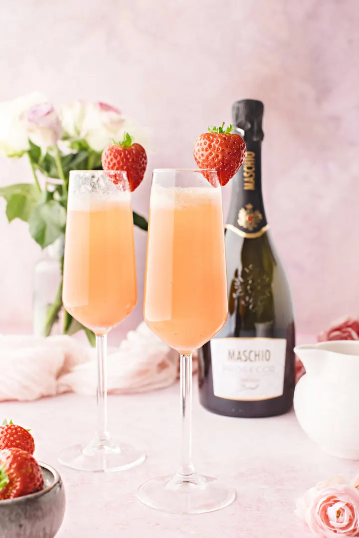 a table set for valentine's day with flowers, champagne flutes of strawberry mimosa with strawberry garnish, and a bottle of champagne