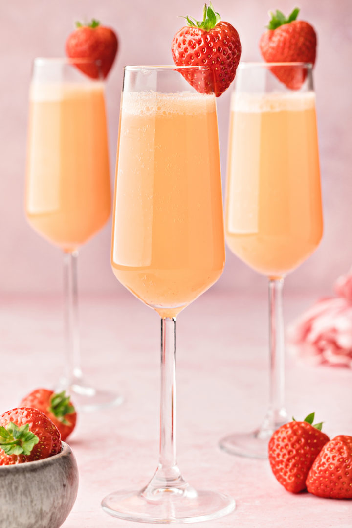 close up photo of 3 champagne flutes filled with a strawberry mimosa on a pink background with a bowl of fresh strawberries