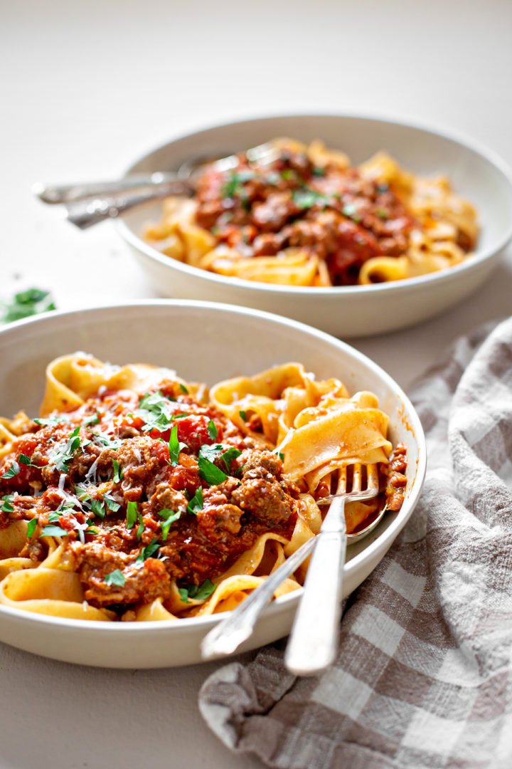 two white pasta bowls on a white surface with tagliatelle pasta and bolognese sauce 
