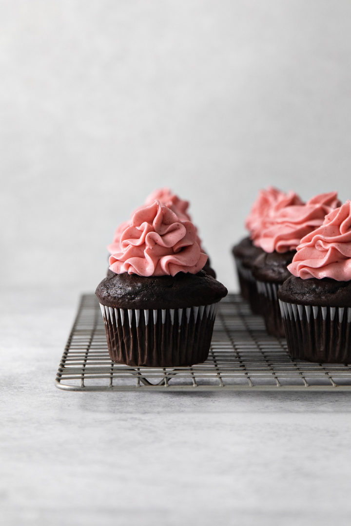 photo of chocolate cupcakes on a cooling rack being frosted with raspberry buttercream