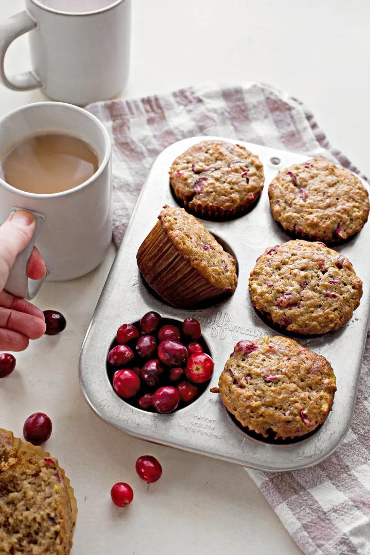woman drinking a cup of coffee next to a muffin tin of this cranberry muffins recipe