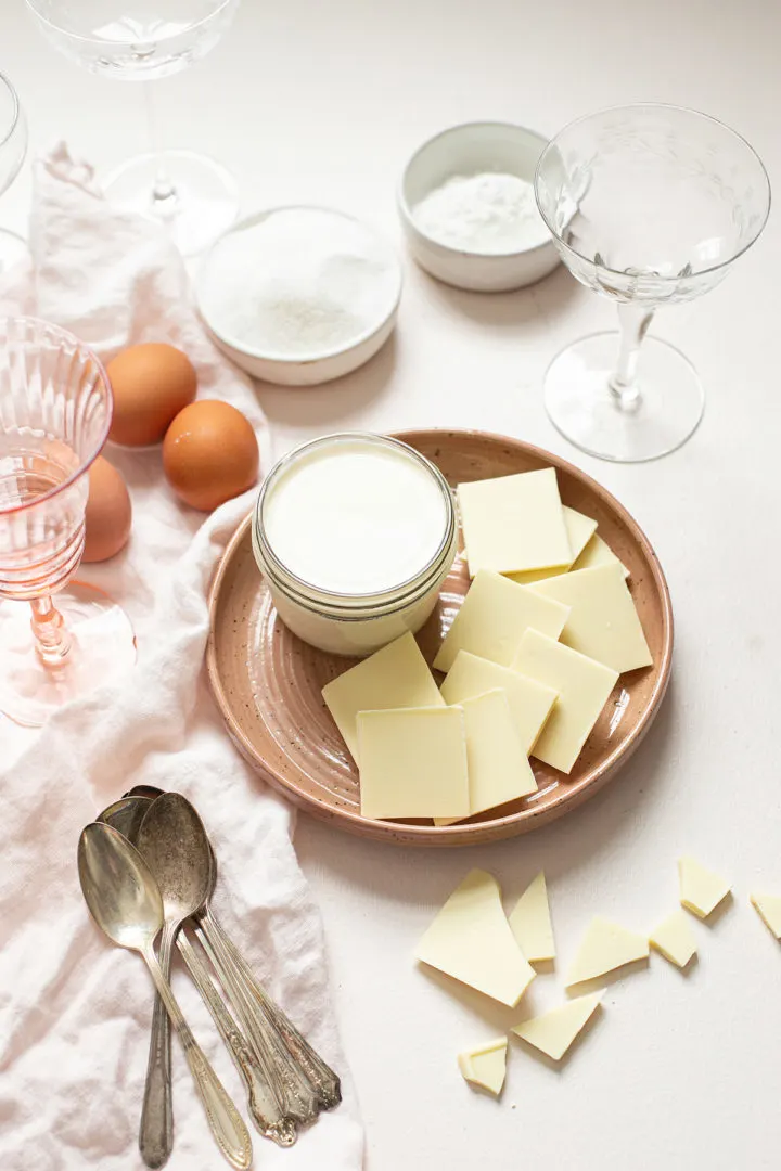 ingredients in this recipe for pudding (white chocolate, milk, sugar, eggs) arranged on a white surfaces with spoons and dessert glasses 