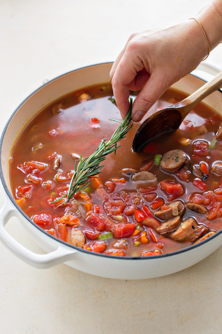 photo of woman adding a sprig of fresh rosemary to a pot of italian soup with sausage