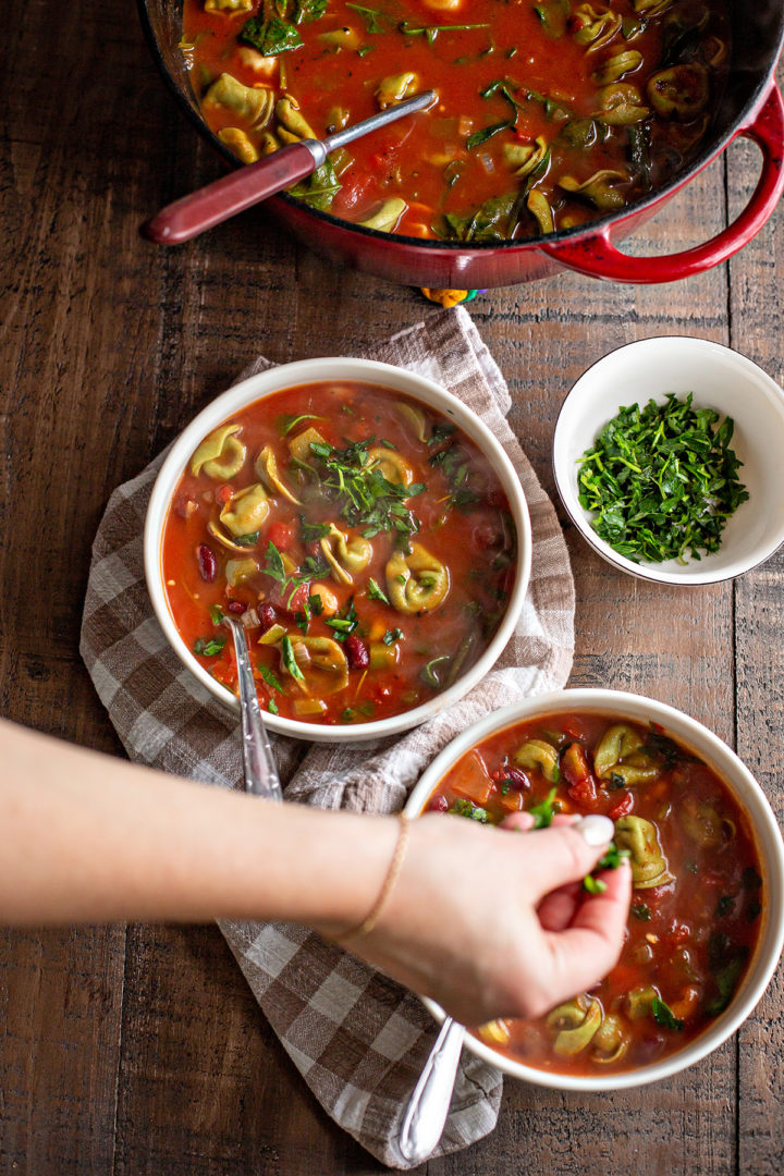 photo of a woman adding fresh herbs on top of bowls of this tortellini soup recipe
