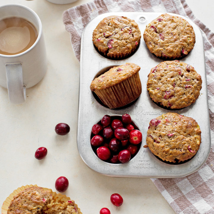 photo of cranberry muffins in a muffin tin next to fresh cranberries and coffee mugs