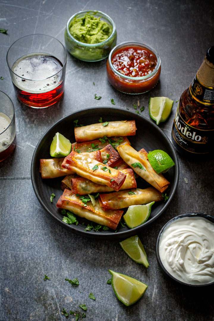 a table set with a plate of baked chicken taquitos, salsa, guacamole, sour cream, and beers for game day appetizers