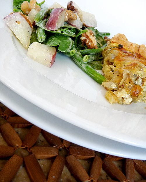 white plate on a wooden placemat plated with a salad and a slice of onion and cheese quiche