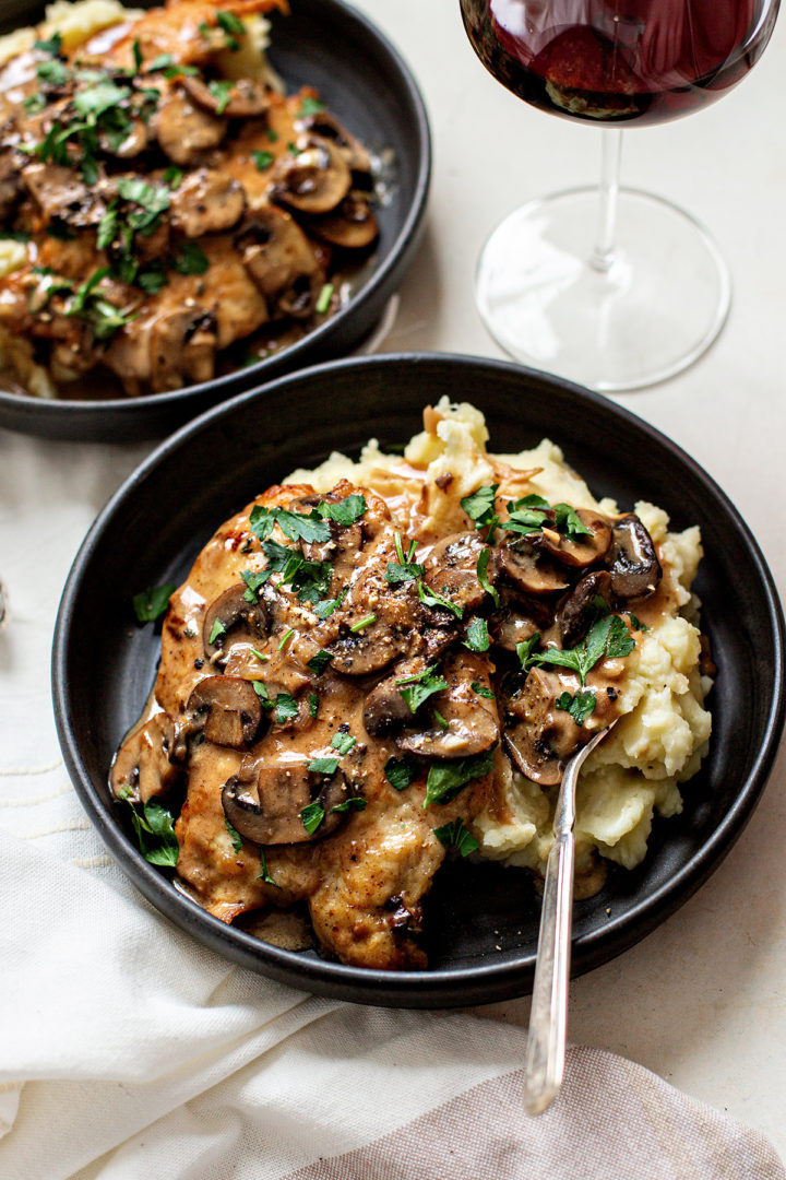 a plate of chicken marsala on a white table with a glass of wine