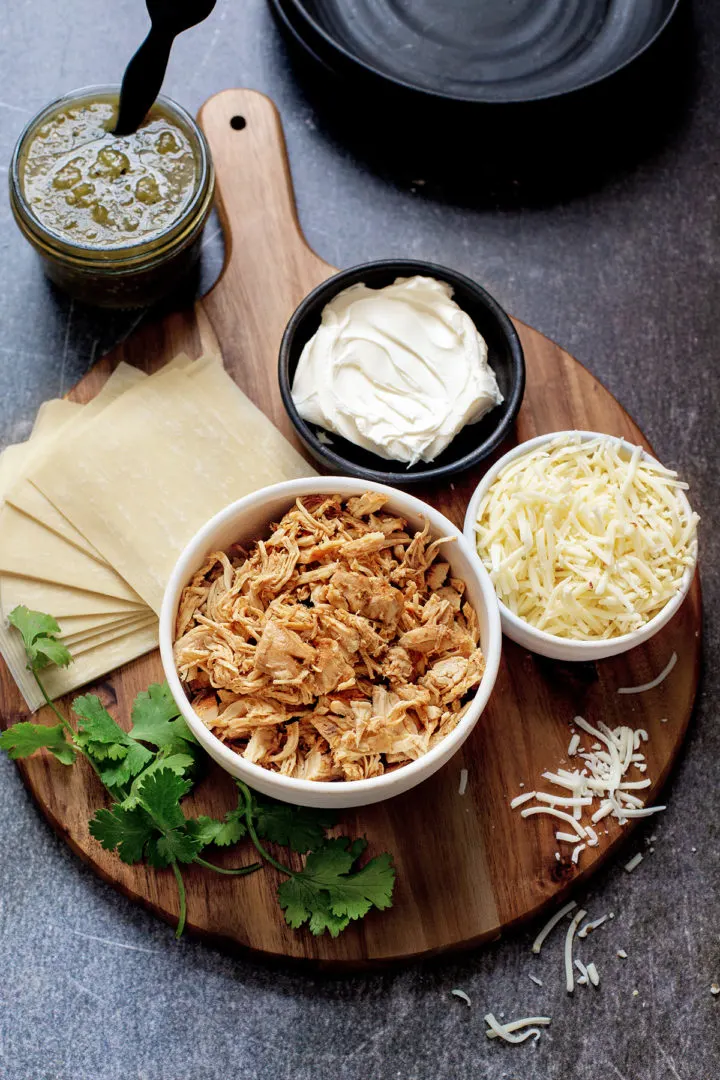 ingredients in this chicken taquitos recipe - chicken, cheese, cilantro, wonton wrappers, cream cheese, and salsa verde arranged on a wooden cutting board