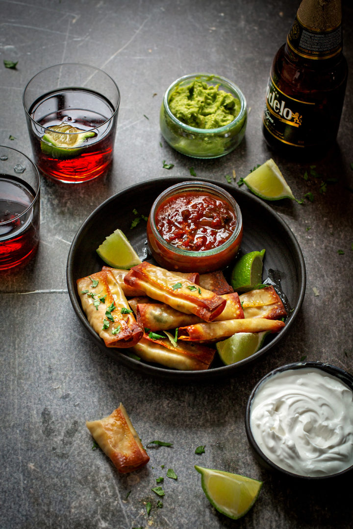 a table set with a plate of cheese and chicken taquitos, bowls of salsa, guacamole, and sour cream, and glasses of beer for game day appetizers