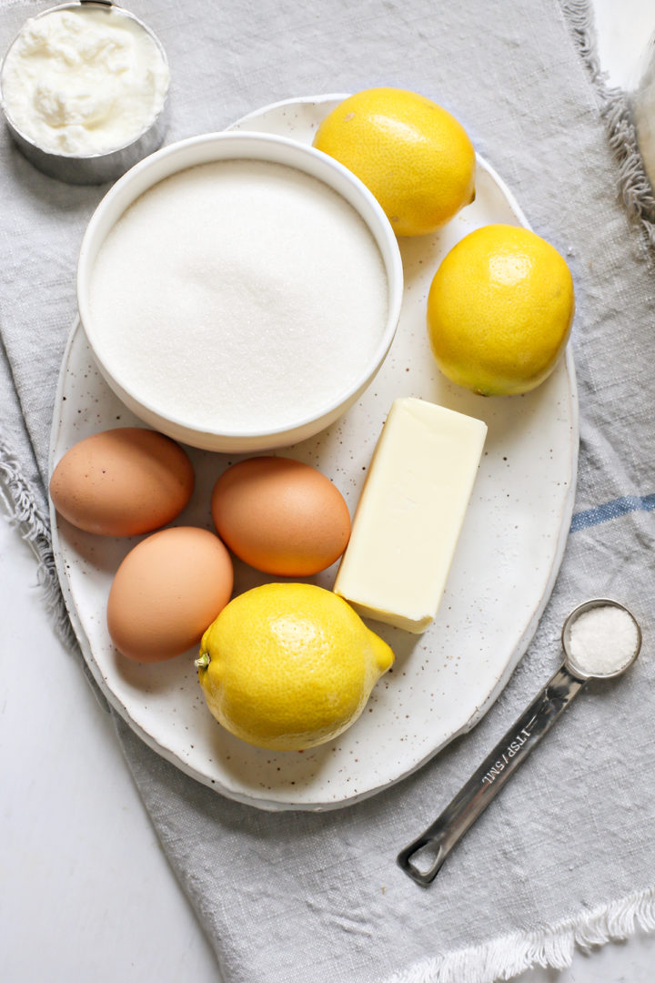 A white oval plate with fresh lemons, butter, eggs, and a bowl of sugar sits atop a grey kitchen towel. Next to the plate is a measuring cup filled with ricotta cheese, showcasing the key ingredients for lemon ricotta bundt cake.