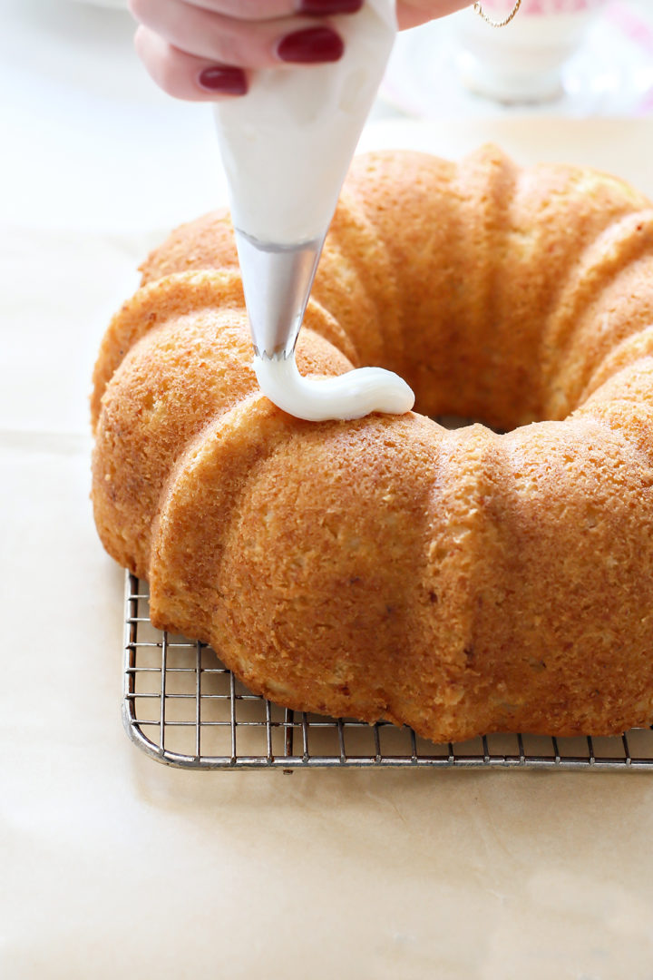 A woman drizzles a smooth lemon glaze over the cooled lemon ricotta bundt cake, which sits on a wire rack over parchment paper.