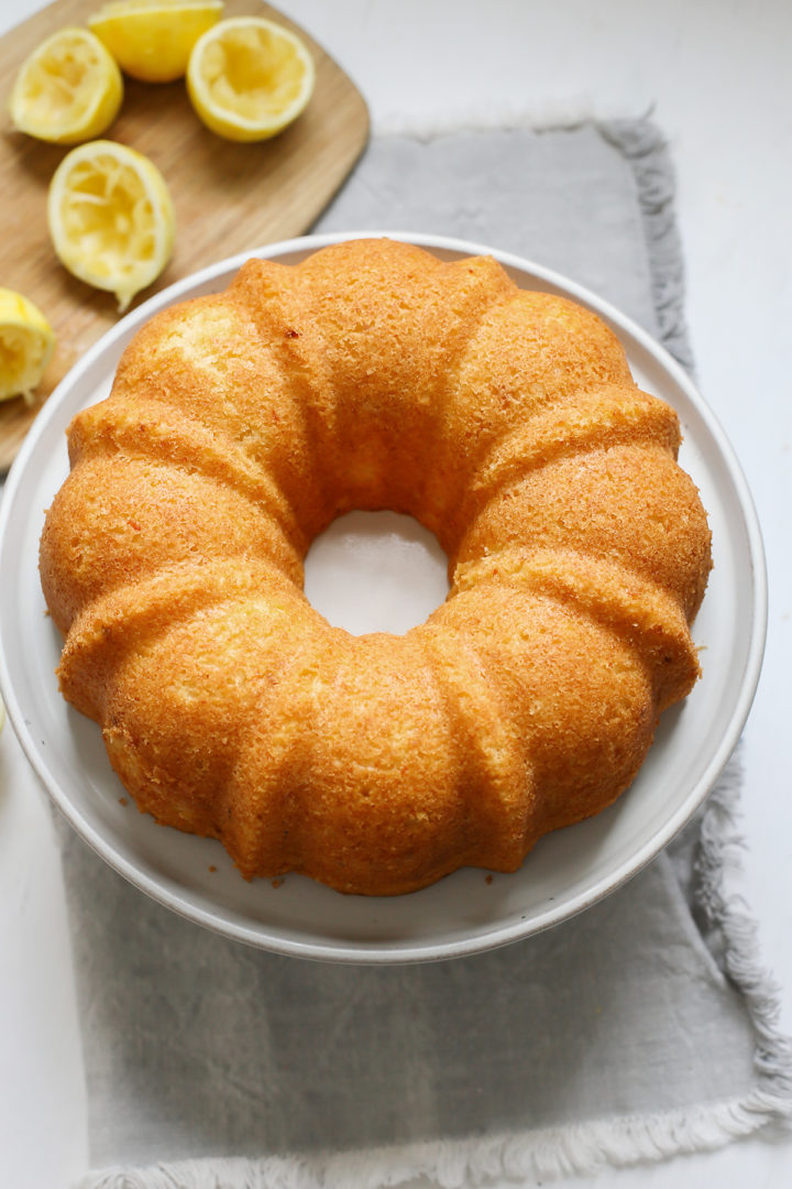 The baked lemon ricotta bundt cake is freshly turned out of the pan and sits on a white cake plate next to a cutting board with freshly squeezed lemons.