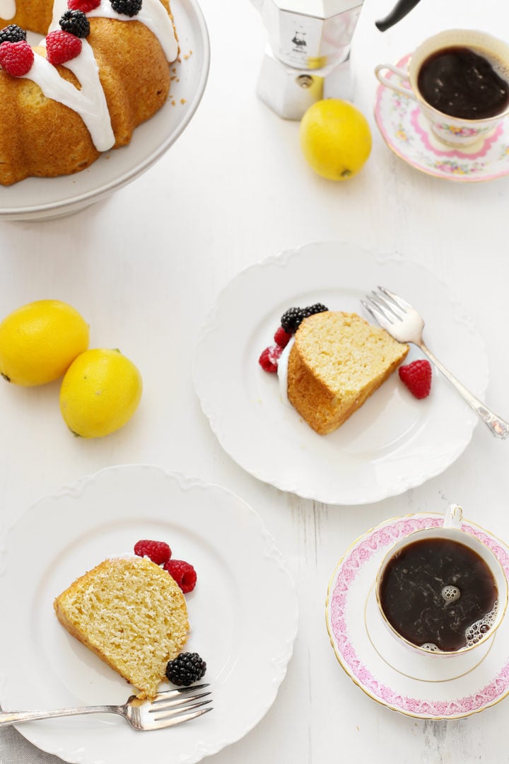 Overhead shot of lemon ricotta bundt cake slices on plates, drizzled with glaze and topped with fresh berries. The scene is set on a white table with fresh lemons and cups of coffee.