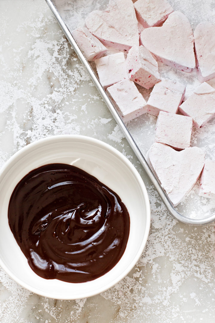 a pan of heart and square homemade marshmallows with raspberries next to a bowl of melted dark chocolate for dipping