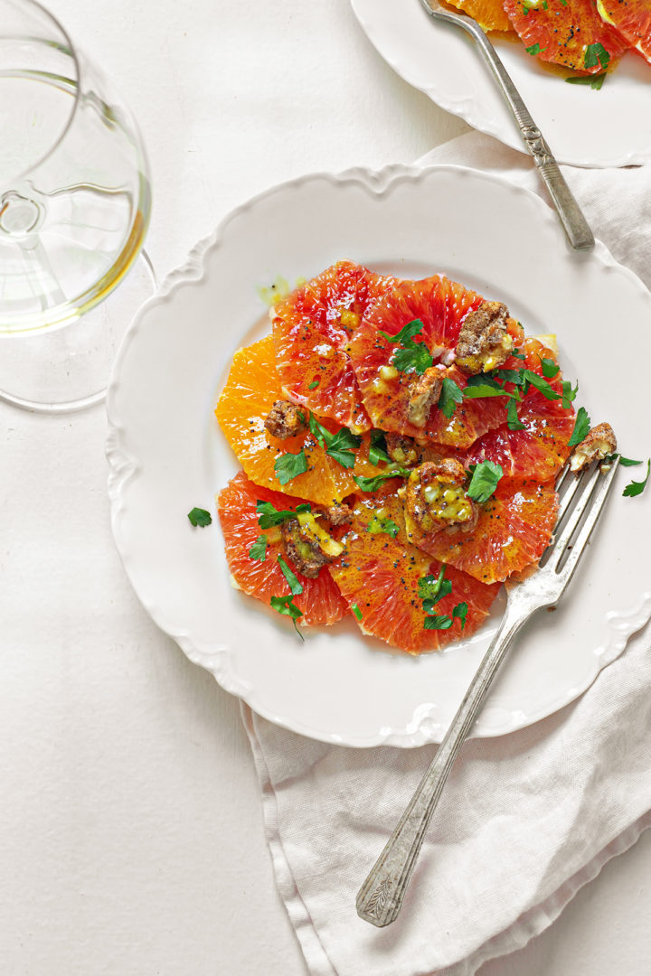 overhead photo of a white plate featuring citrus salad next to a wine glass on a white table