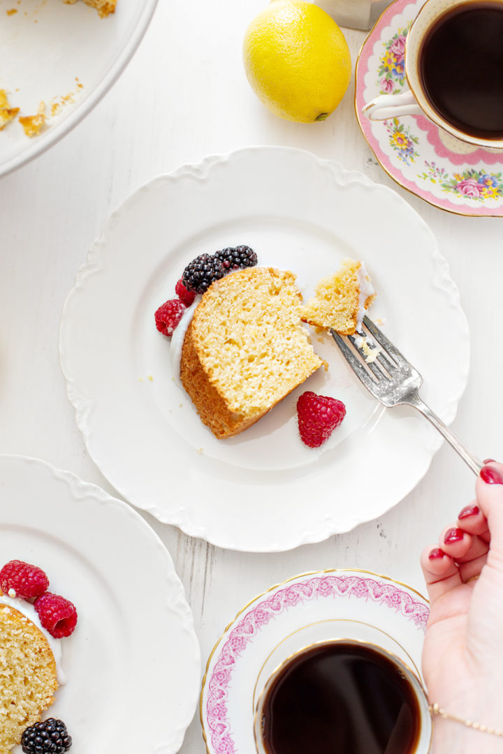 Close-up shot of a plated lemon ricotta bundt cake slice with glaze and fresh berries. A woman is cutting into the cake with a fork, revealing its moist and tender texture.