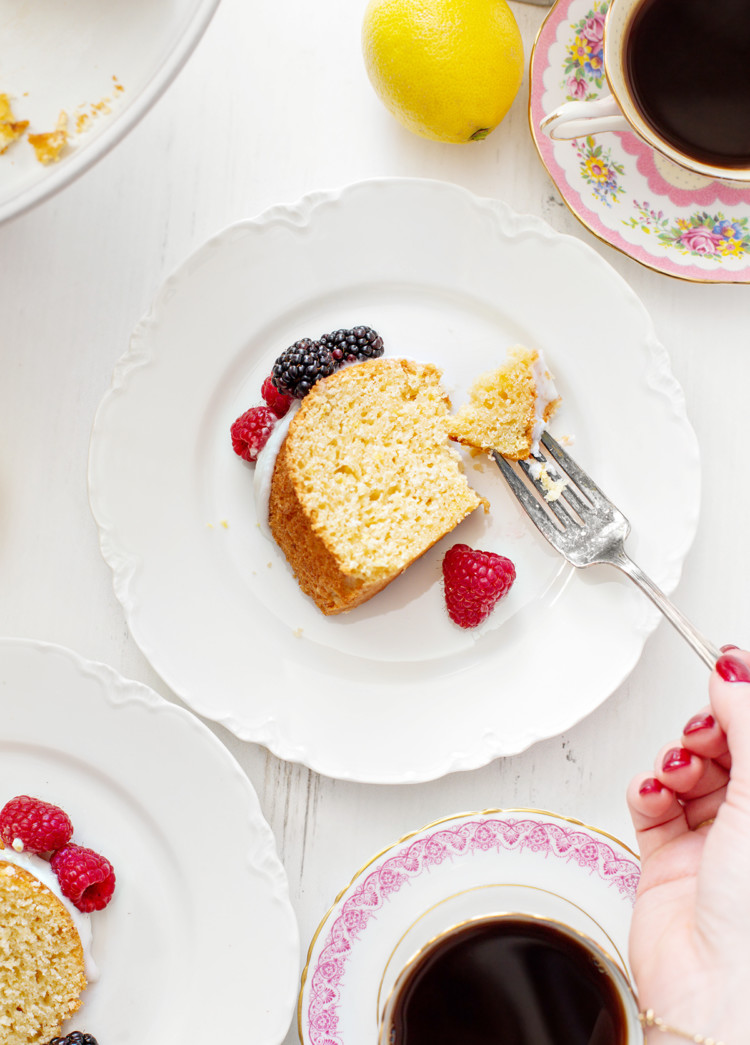 Close-up shot of a plated lemon ricotta bundt cake slice with glaze and fresh berries. A woman is cutting into the cake with a fork, revealing its moist and tender texture.
