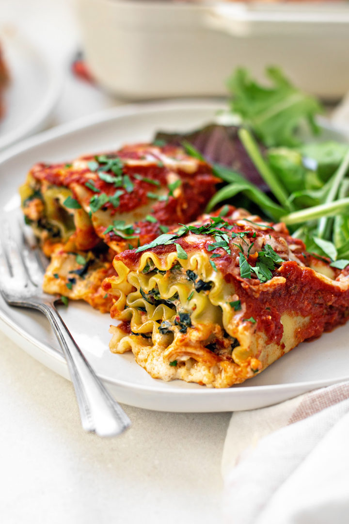 photo of two spinach lasagna rolls on a white plate with salad and a fork