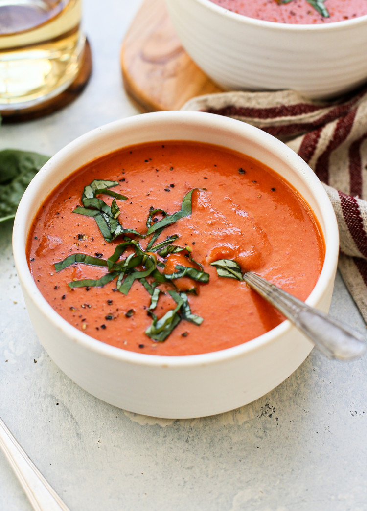 photo of a white bowl of cream of tomato soup with a spoon and fresh basil on a light blue background