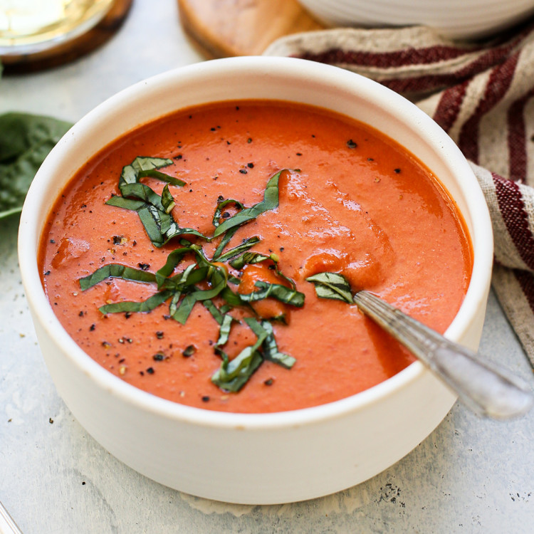 photo of a white bowl of cream of tomato soup with a spoon and fresh basil on a light blue background