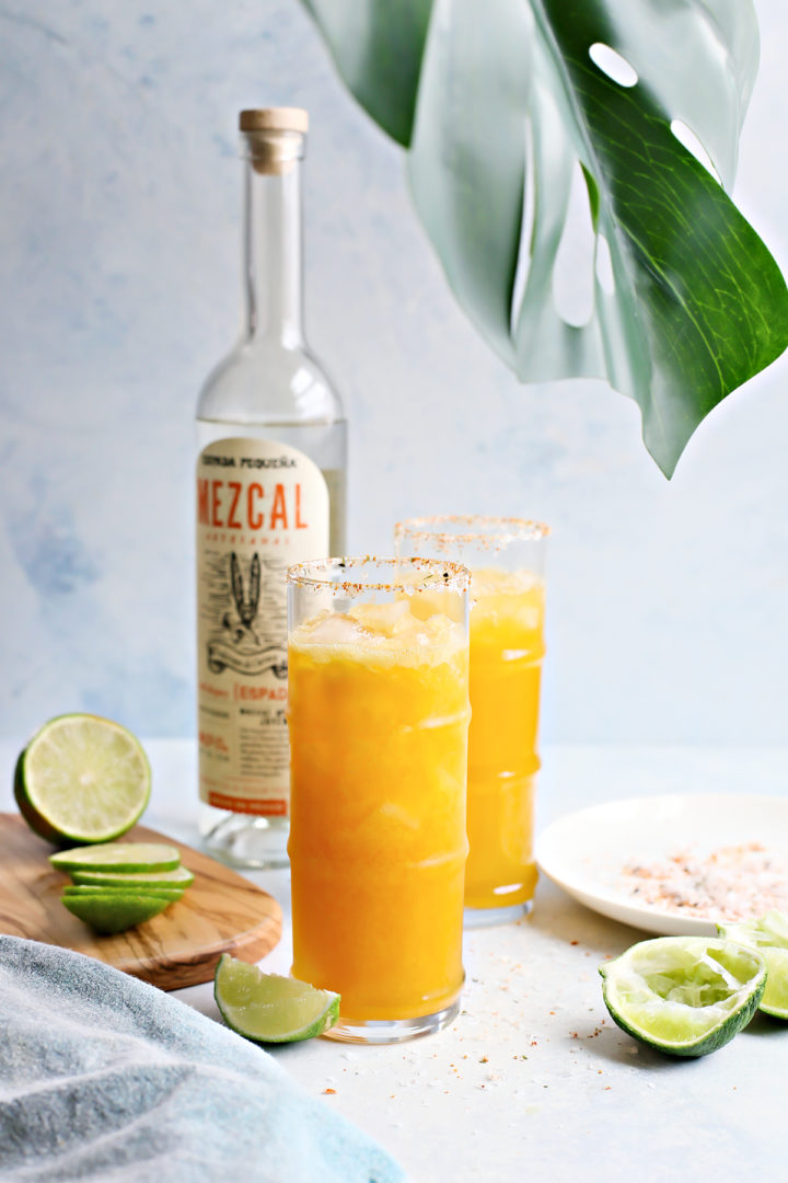 two glasses of passion fruit margaritas next to a plant, fresh limes, and bottle of mezcal