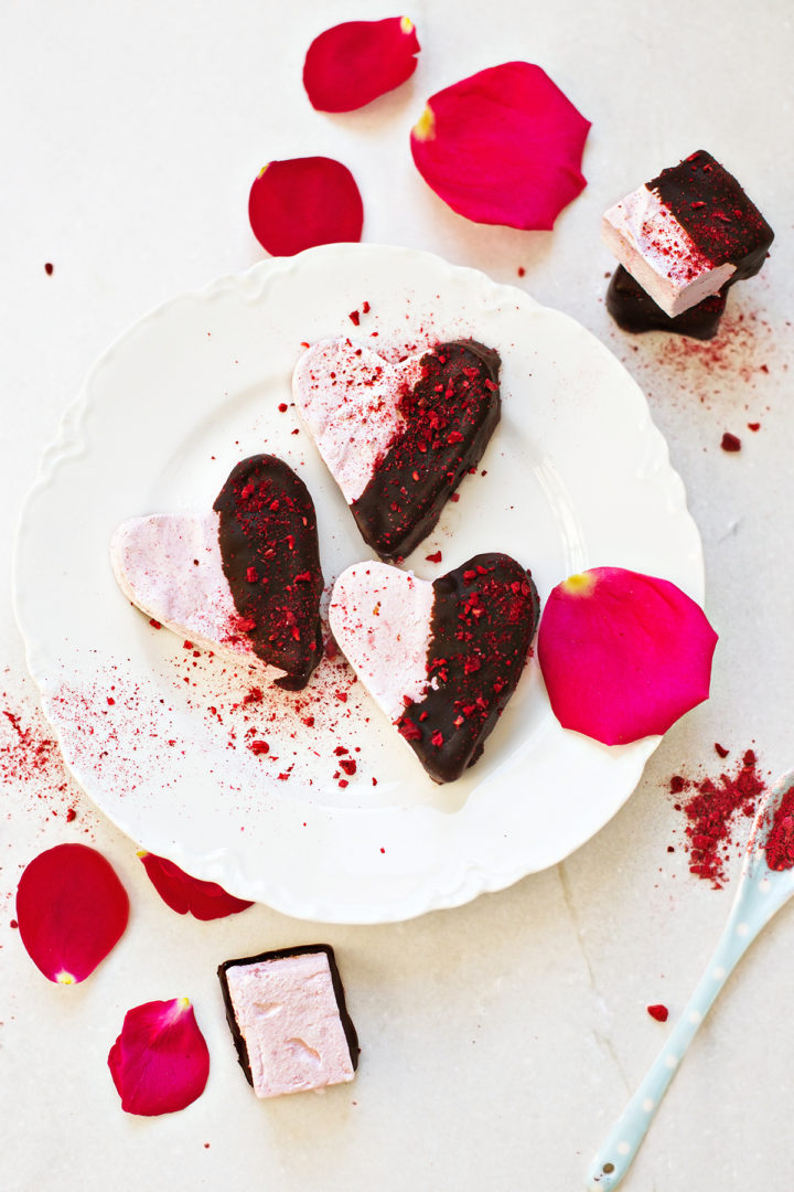 a white plate with three chocolate covered raspberry marshmallows on it, on a white background surrounded by rose petals