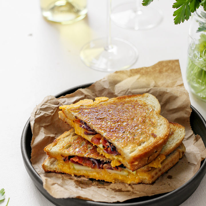 two slices of apple and bacon grilled cheese on a plate served with a glass of white wine