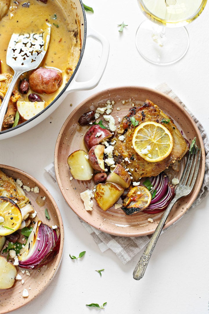 greek chicken thighs being served from a cast iron skillet onto plates on a white table