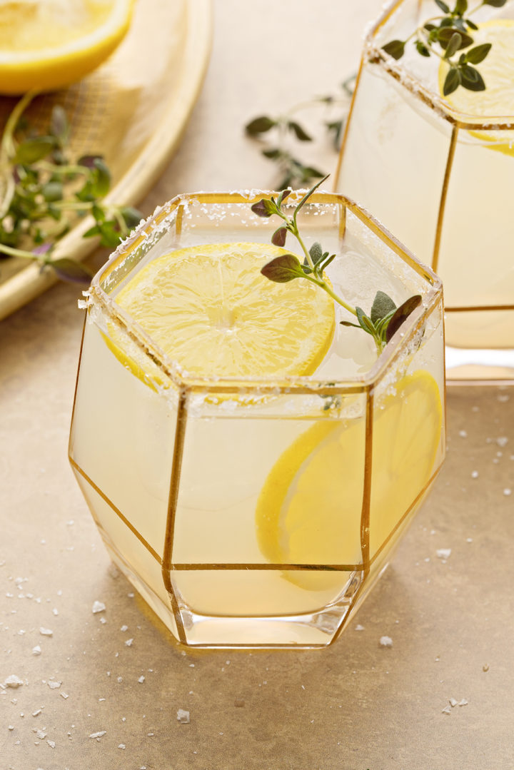 photo of a lemon margarita on a gold background, garnished with lemon slices and fresh thyme