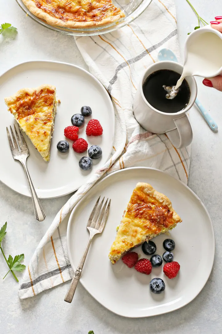 overhead photo of a plate of lorraine quiche next to a mug of coffee, a woman pouring cream into the coffee mug