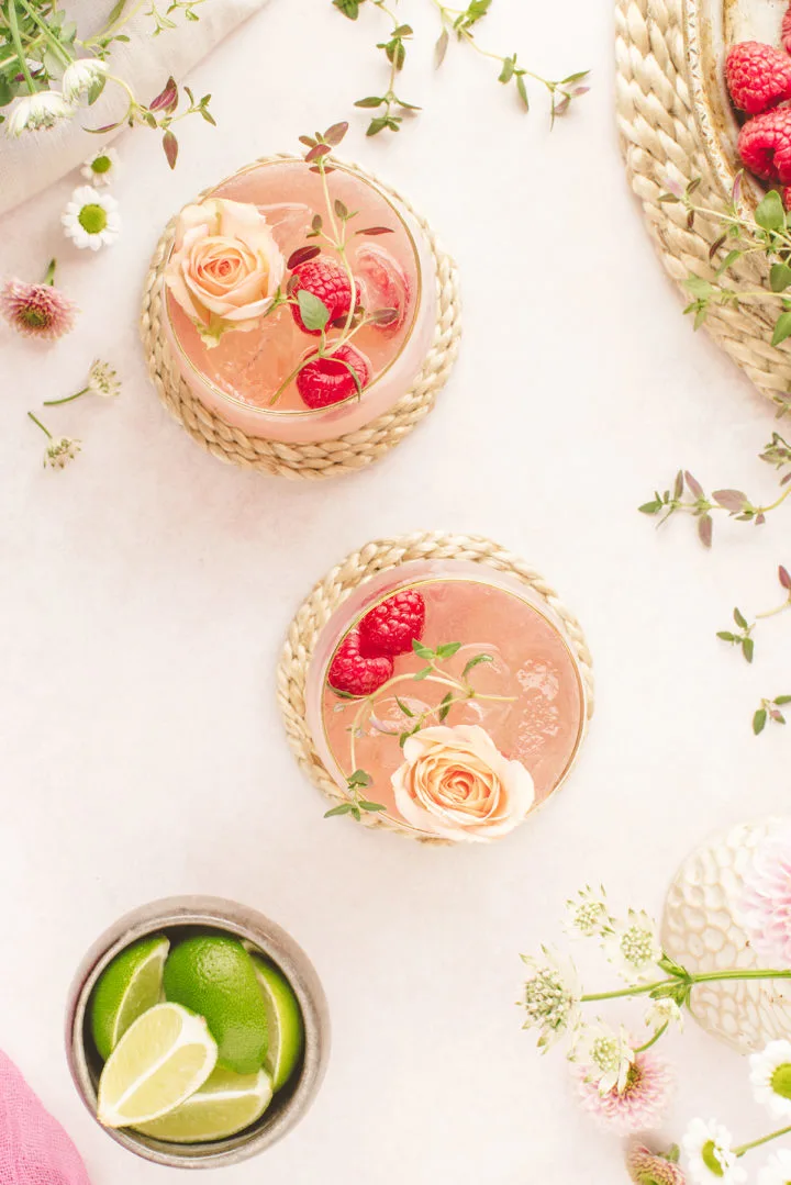 overhead photo of two glasses of a floradora cocktail (a raspberry gin cocktail for summer) with fresh raspberries, flower garnishes, and a bowl of  sliced fresh limes