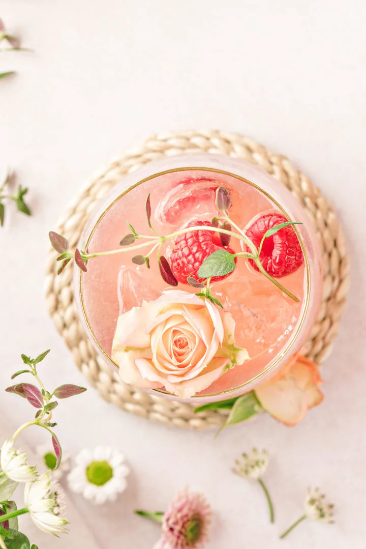 overhead photo of a floradora (a gin cocktail recipe) in a glass garnished with fresh thyme, raspberries, and edible roses