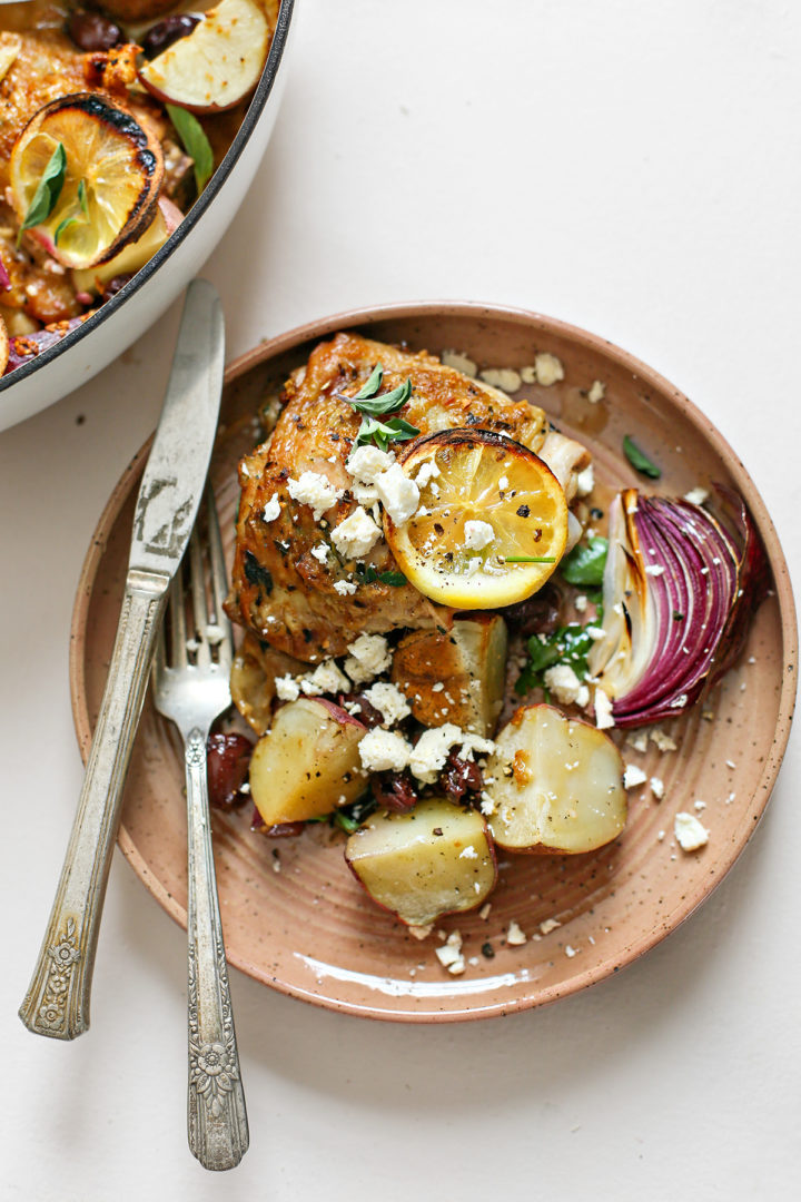 overhead photo of greek chicken thighs on a plate with potatoes, lemons, olives, and feta