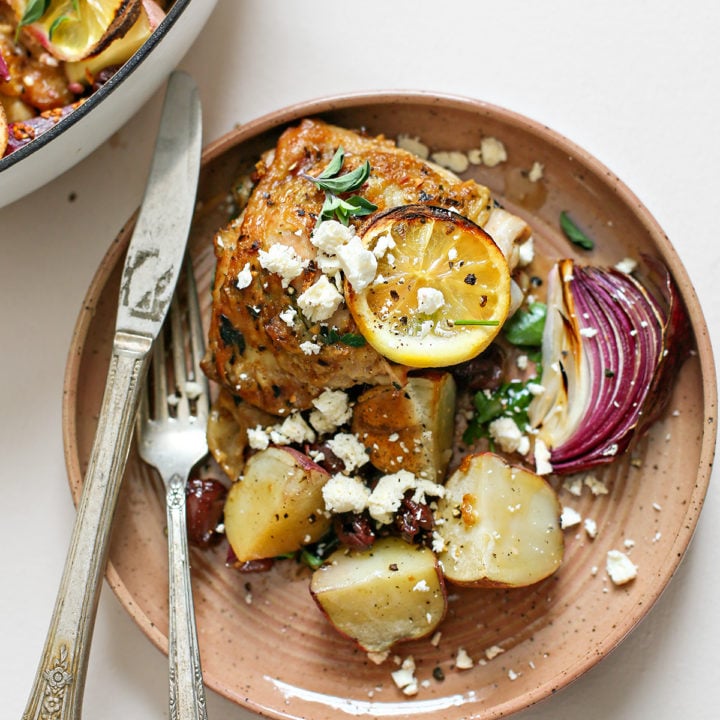 overhead photo of greek chicken thighs on a plate with potatoes, lemons, olives, and feta