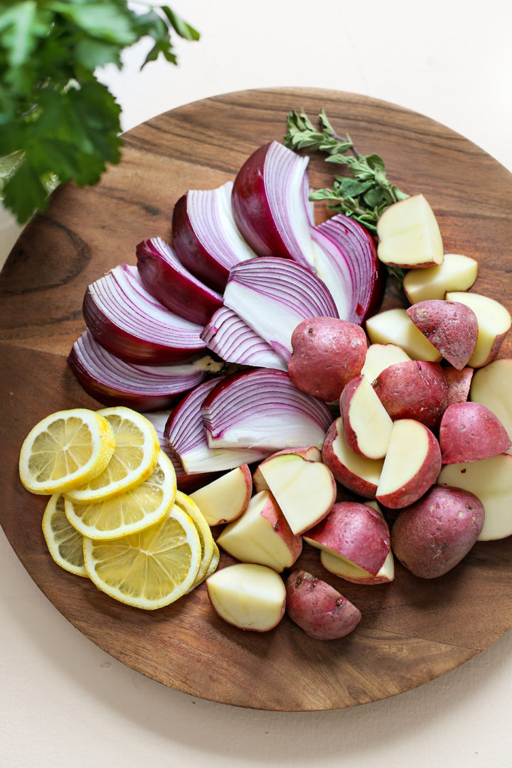 cut potatoes, red onions, sliced lemon, and fresh oregano on a wooden cutting board to make skillet chicken thighs
