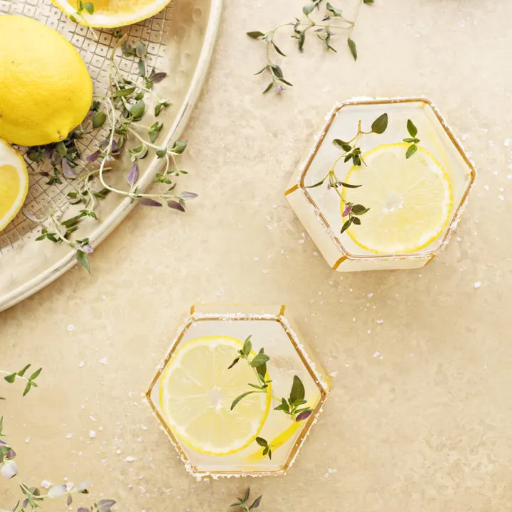 overhead photo of two lemon margaritas on a gold background with fresh thyme and lemon