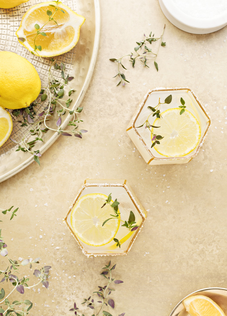 overhead photo of two lemon margaritas on a gold background with fresh thyme and lemon