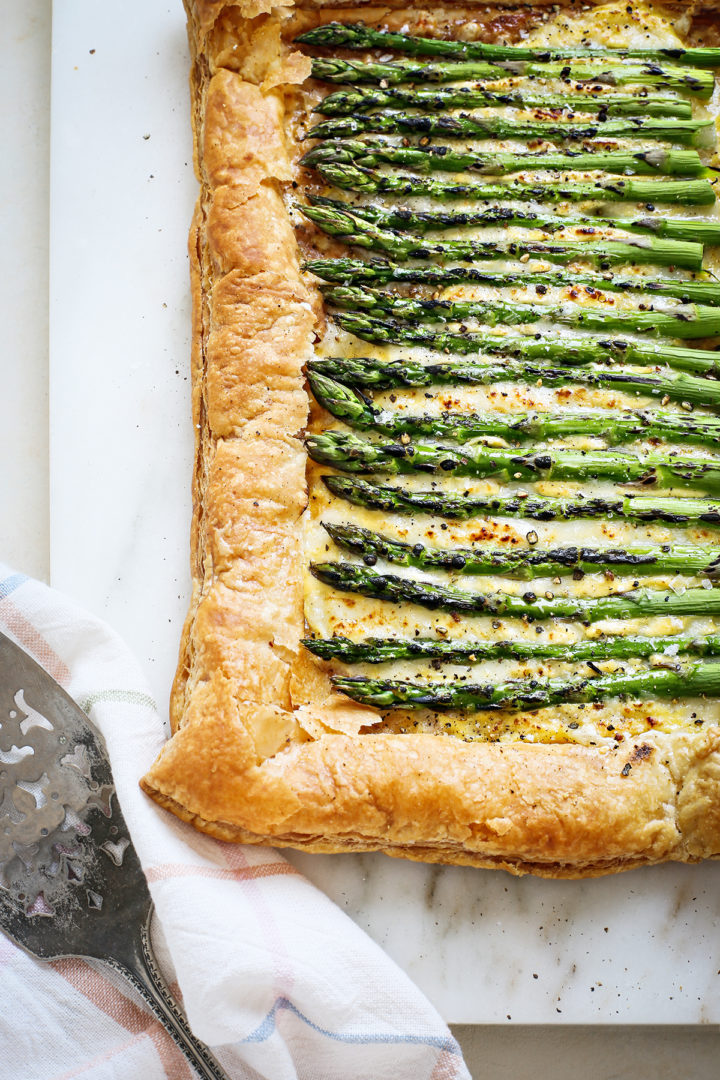 an asparagus puff pastry tart on a white marble board with a serving spatula