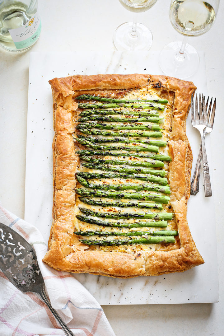 asparagus tart with puff pastry on a white marble cutting board with forks and glasses of white wine