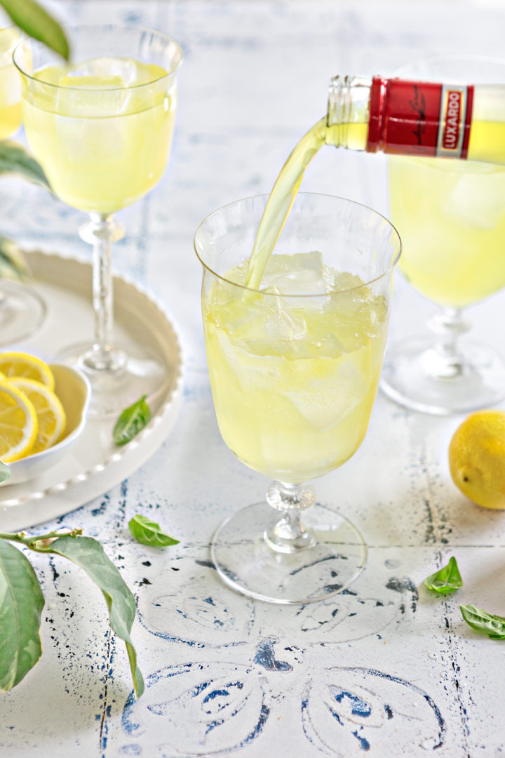 limoncello being poured into a glass with ice and prosecco. also in the photo are prepared limoncello spritzers, fresh lemons, and basil leaves
