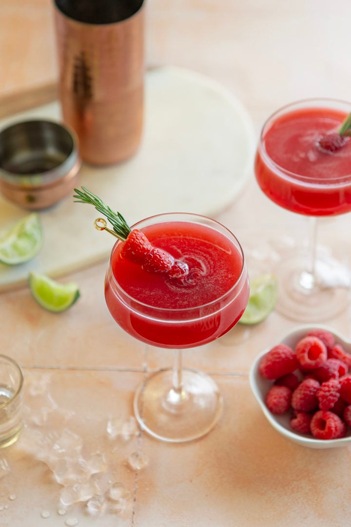 two glasses of a raspberry cosmopolitan next to a bowl of raspberries and slices of lime