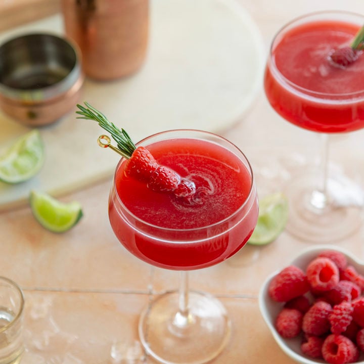 two glasses of a raspberry cosmopolitan next to a bowl of raspberries and slices of lime