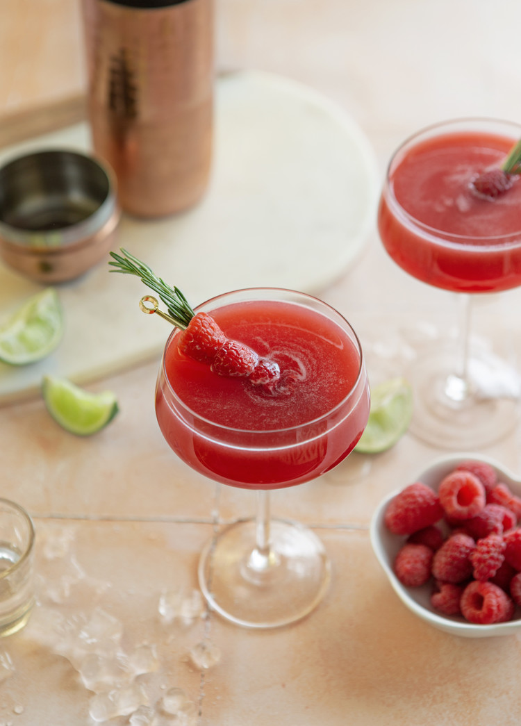 two glasses of a raspberry cosmopolitan next to a bowl of raspberries and slices of lime