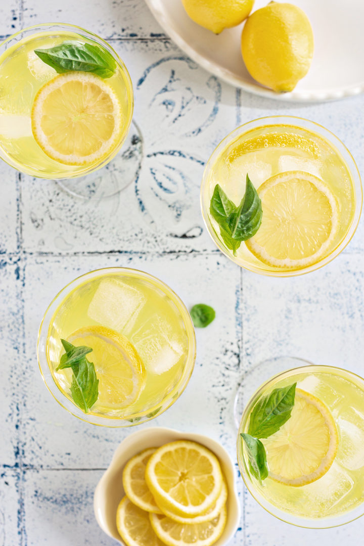 An overhead view of a multiple limoncello spritzers in a large wine glasses, surrounded by fresh lemons and a bowl of sliced lemons on a white and blue tile background