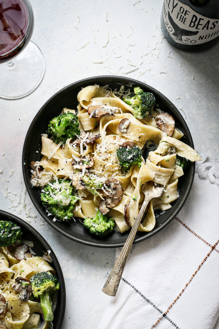 two bowls of mushroom alfredo next to a glass of wine