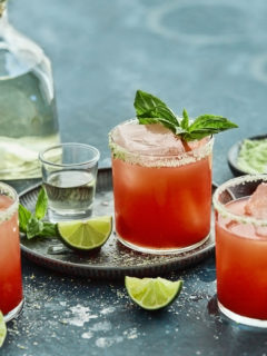 3 glasses of strawberry basil margarita on a teal background with a bottle of tequila, fresh lime wedges and basil sprigs
