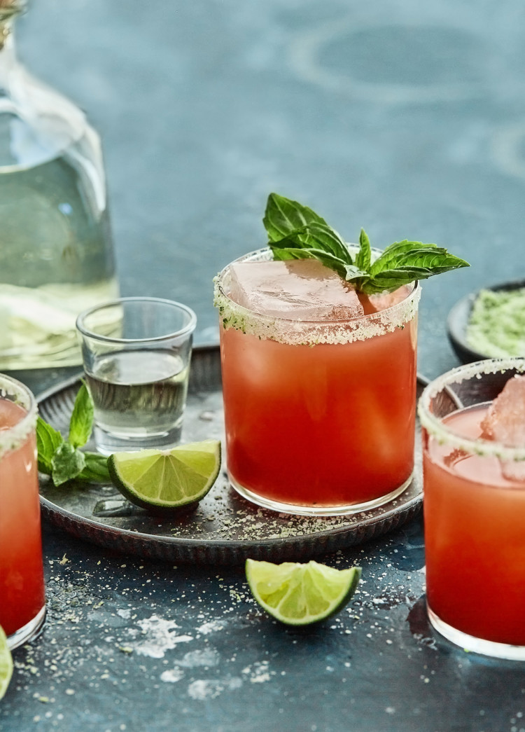 3 glasses of strawberry basil margarita on a teal background with a bottle of tequila, fresh lime wedges and basil sprigs