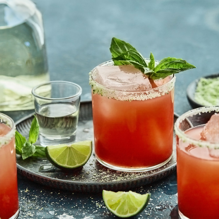 3 glasses of strawberry basil margarita on a teal background with a bottle of tequila, fresh lime wedges and basil sprigs